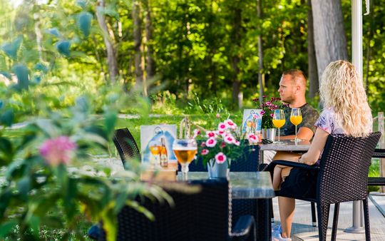 Een stel zit buiten op een terras wat te drinken, ze zijn omgeven door een groene omgeving. Bij het Proeflab in het Waterloopbos in de Noordoostpolder.