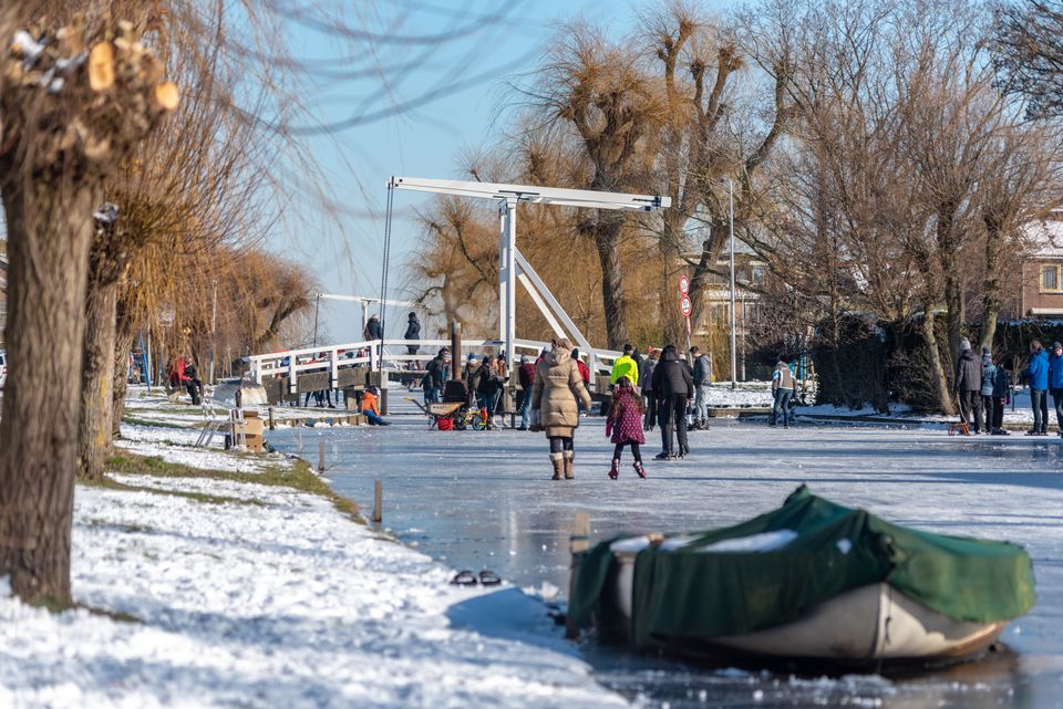 Een winterse dag in Alphen aan den Rijn: bewoners en bezoekers genieten van het ijs op een bevroren sloot, terwijl schaatsers en wandelaars elkaar ontmoeten bij de karakteristieke ophaalbrug. De besneeuwde oevers, kale bomen en heldere blauwe lucht geven het tafereel een rustige, bijna nostalgische sfeer waarin samen buiten zijn centraal staat.