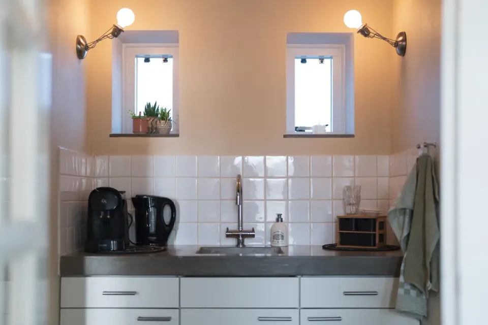Een wit keukenblokje met witte tegeltjes, onder een oranje muur met 2 kleine raampjes. Koffie en theevoorzieningen en handdoekjes.