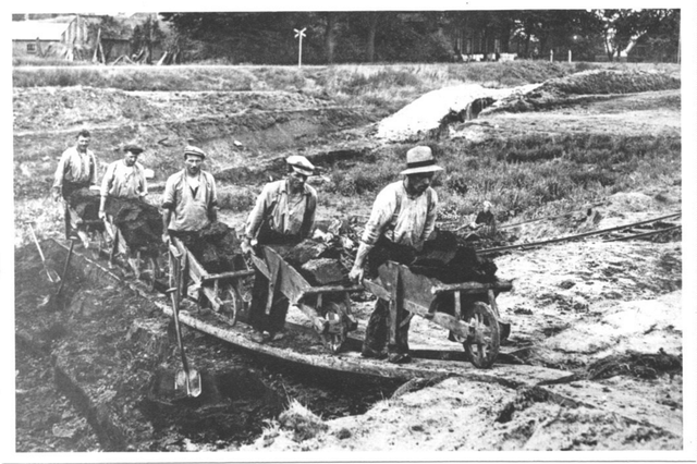 Zwart-wit foto van veenarbeiders die met kruiwagens vol turf over een plank lopen.