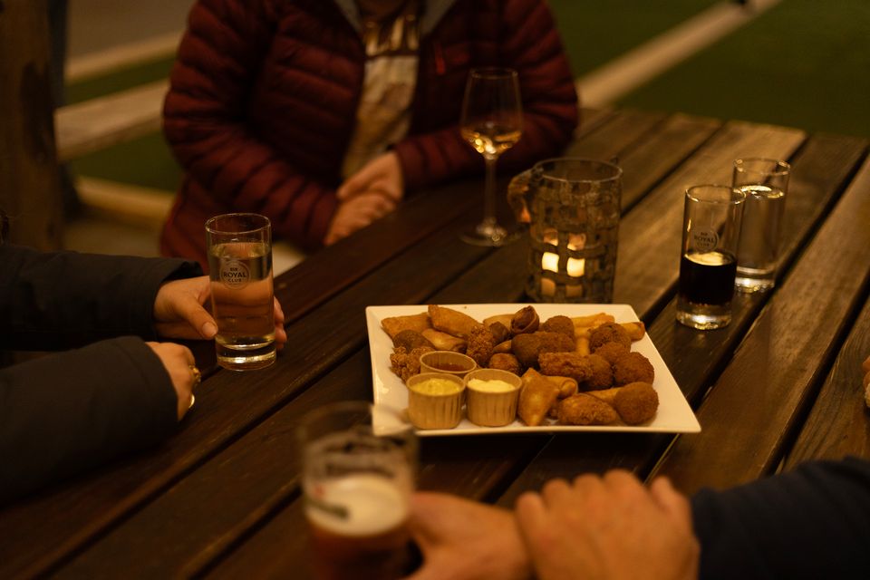Gezellig borrelmoment op een houten terras met warme sfeerverlichting, waar een groep mensen geniet van een royale schaal bittergarnituur met sauzen en diverse drankjes