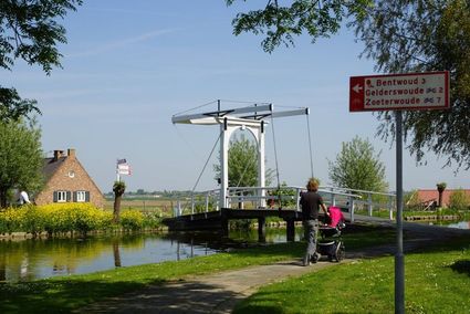 Wandelaars met kinderwagen bij een wit ophaalbruggetje over het water in de groene omgeving van Gelderswoude, met zicht op weilanden en bewegwijzering naar omliggende dorpen.