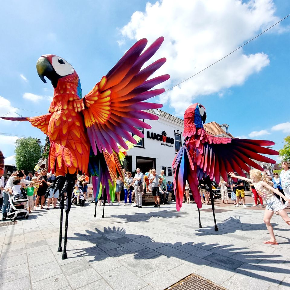 Grote kleurrijke papegaaien op stelten tijdens een straattheaterfestival in het centrum van Woerden, terwijl kinderen en bezoekers toekijken.