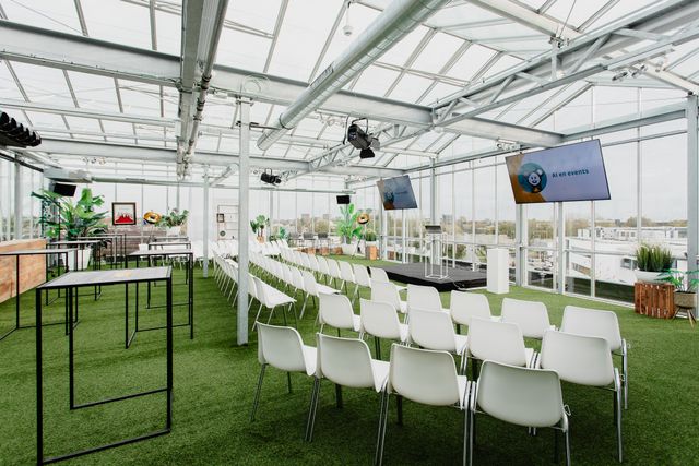 Binnenaanzicht van KASXL, een moderne en lichte vergader- en evenementenlocatie van KAS Meeting & Eventlocatie. De ruimte is ingericht met rijen witte stoelen gericht op een podium met schermen waarop “AI en events” staat. De locatie heeft een glazen kasconstructie, kunstgrasvloer, hoge tafels, veel daglicht en is omgeven door planten en stadsuitzicht.