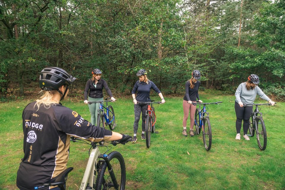 Vier personen staan met een mountainbike te luisteren naar persoon die uitlegt geeft. In het bos voor een mountainbikepad.