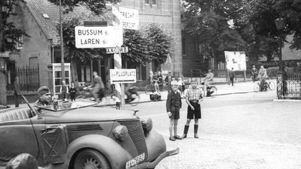Zwart wit foto van kruispunt met auto van Wehrmacht en twee jongetjes die toekijken