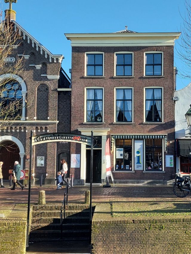 Toeristisch informatiepunt TIP Oudewater, Groene Hart. Historisch grachtenpand aan de gracht met bezoekersinformatie, naast een monumentale kerk en brug, terwijl wandelaars passeren in de binnenstad op een zonnige dag.