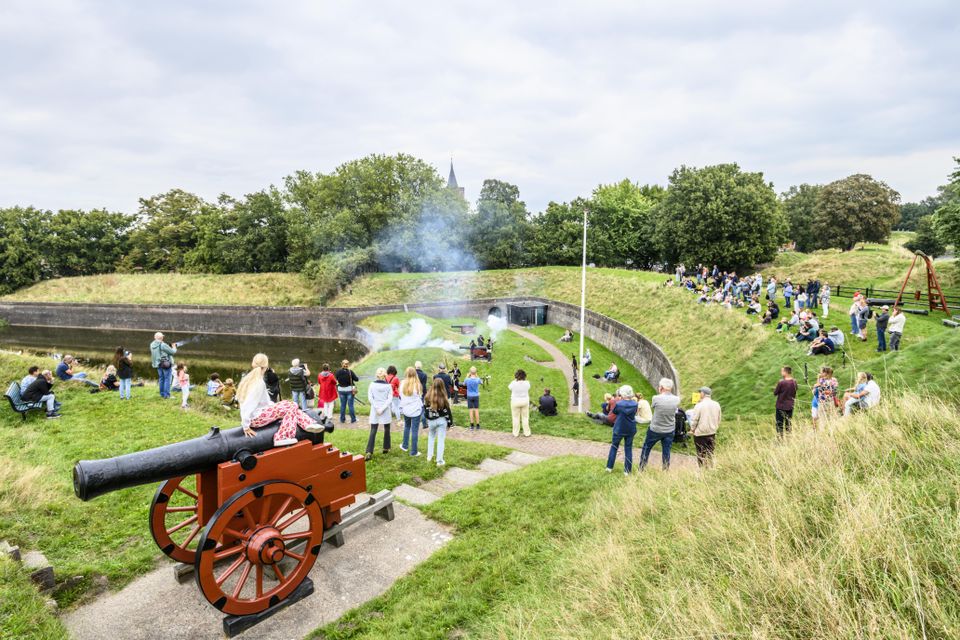 Grote groep mensen kijkt vanaf de vestingwallen van Naarden naar een voorstelling tijdens de Schuttersdag in het Nederlands Vestingmuseum. Op de voorgrond zie je een meisje dat op een historisch kanon zit en naar het schouwspel kijkt.