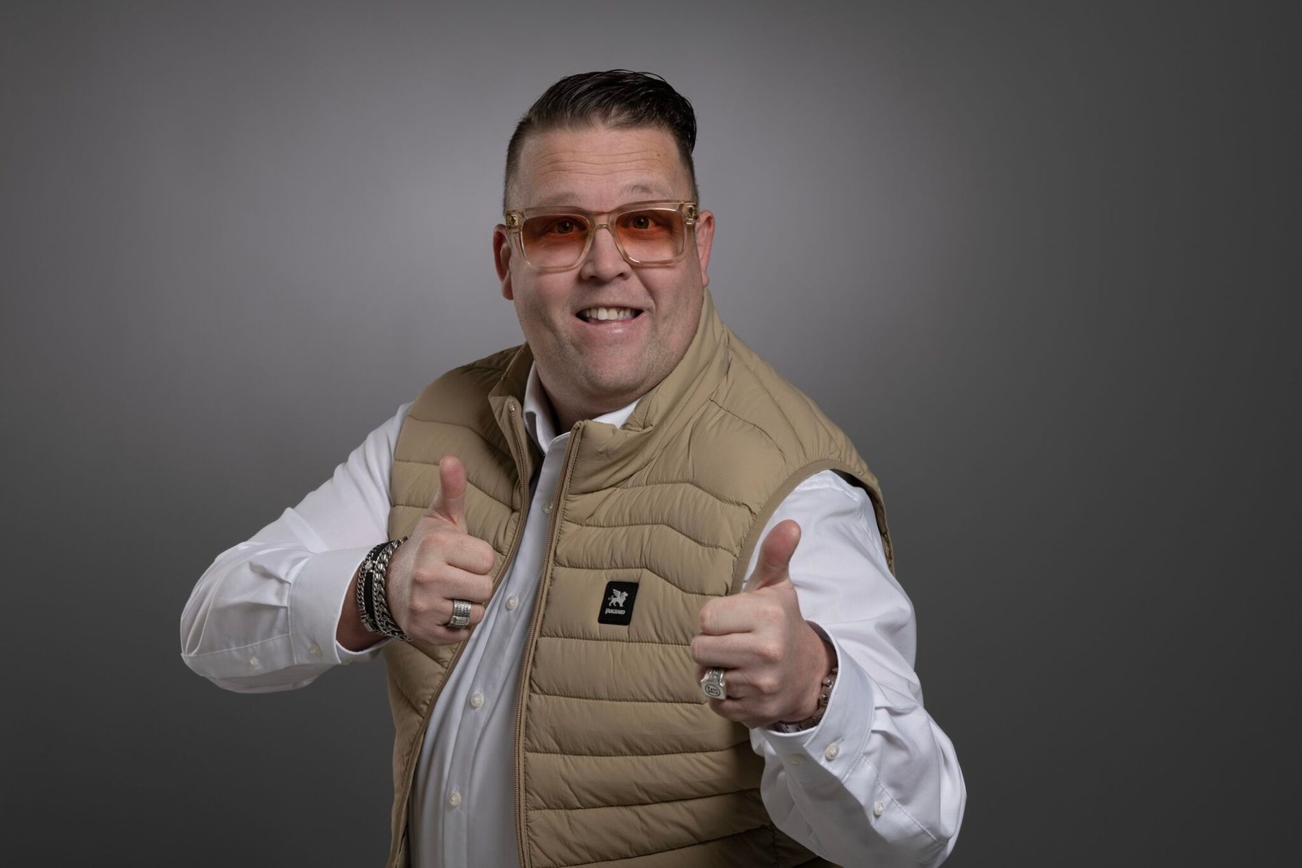 Foto van een artiest in wit overhemd met beige bodywarmer en zonnebril op tegen een grijze achtergrond.