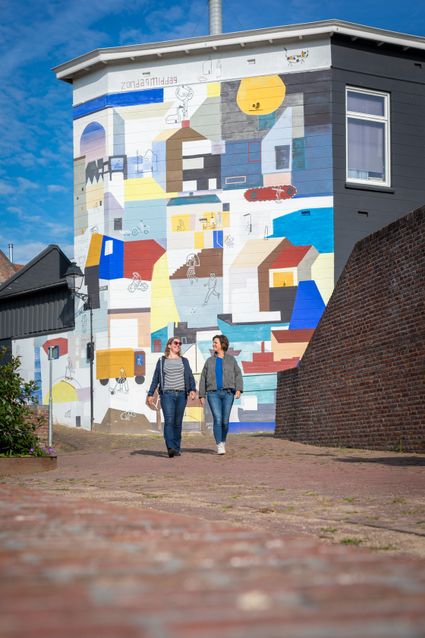 Twee personen die samen door een straat lopen langs een gebouw met een grote kleurrijke muurschildering, bestaande uit abstracte vormen en stadsdetails, op een zonnige dag in een stedelijke omgeving.