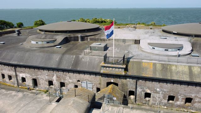 Een betonnen forteiland gelegen in het water van het IJmeer, gefotografeerd vanuit de lucht. Bovenop het fort wappert de Nederlandse vlag.