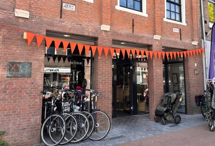 Voorgevel van kringloopwinkel De Terugwinning in het centrum van Woerden, met oranje vlaggetjes, fietsen voor de deur en kledingrekken buiten op straat.