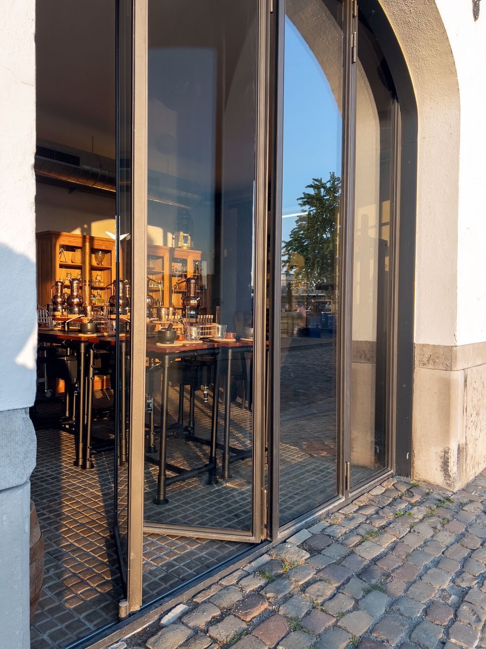 Ingang van Het Stookatelier in Maastricht aan de Oeverwal 8, met open glazen deuren en een blik naar binnen op de ambachtelijke werktafels en koperen distilleerketels. De avondzon werpt warme reflecties op de cobblestones en glazen pui.