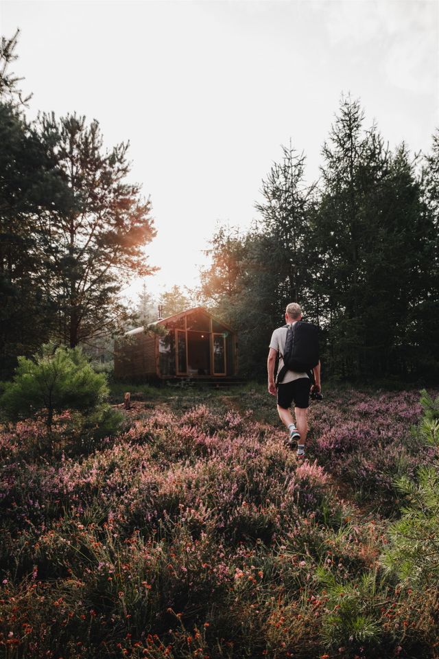 Een man loopt met zijn backpack op door paarse heide richting een cabin.