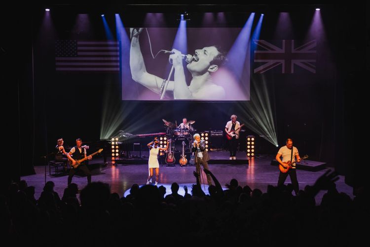 Foto van een podium met een 7-koppige band en instrumenten. Boven hen wordt Freddy Mercury getoond op een scherm.