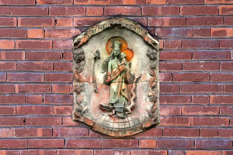Een muur waar een plaquette op is bevestigd. Maria met kind staande op een wereldbol en draak
