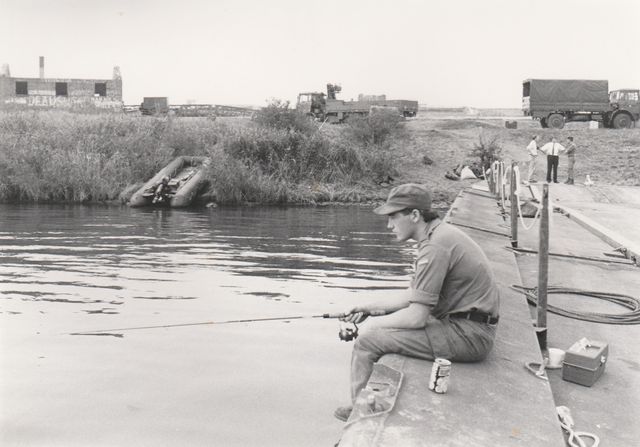Een visser zit op de pier van Pampus. Op de achtergrond zijn militaire wagens te zien.