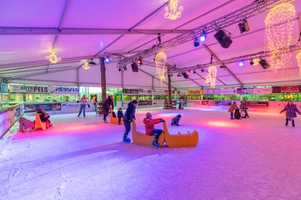 Een kleurrijk en sfeervol beeld van de overdekte ijsbaan in Woerden, waar kinderen en ouders samen genieten van een middag schaatsplezier. De warme lichtkleuren, speelse hulpmiddelen en fonkelende decoratie creëren een vrolijke, winterse ambiance waarin plezier en beweging centraal staan.