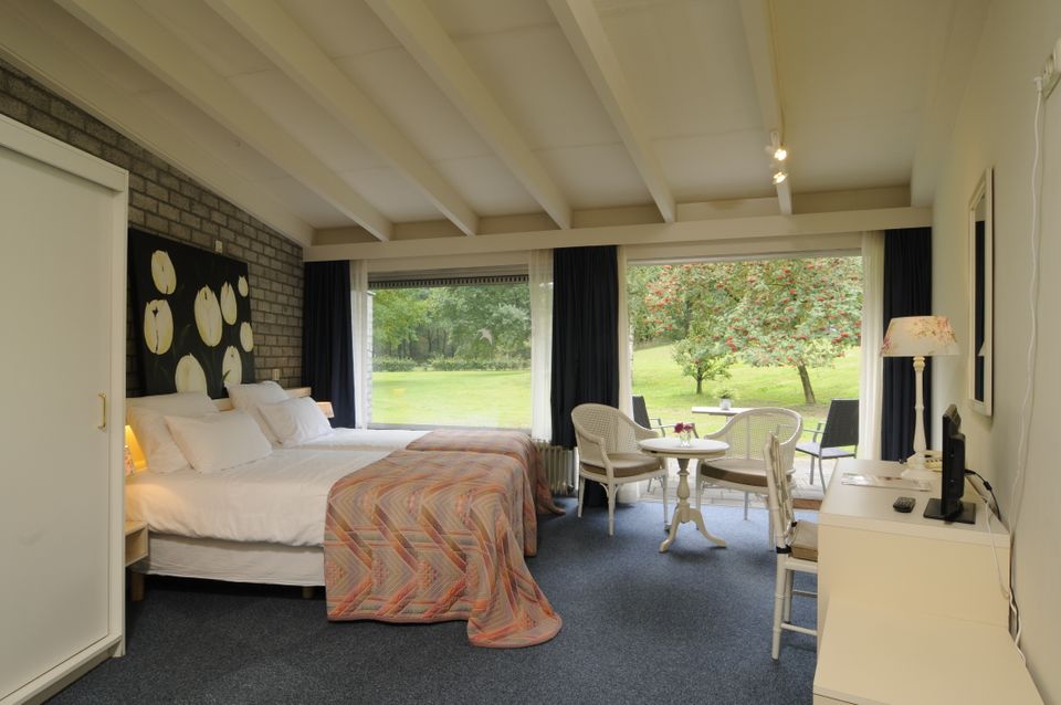 Hotelkamer met grote ramen en uitzicht op een tuin. Een groot bed midden in de kamer met blauwe vloerbedekking en donkere achterwand.