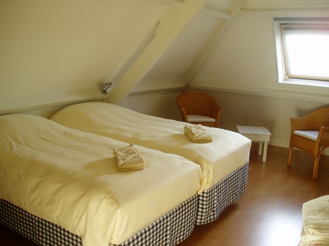 Slaapkamer onder de schuine kap met twee 1-persoonsbedden. Aan de rechterkant zit een dakraam met daaronder twee rotan stoelen en een tafeltje.