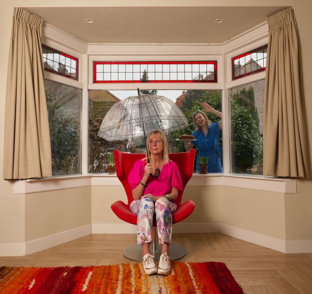 dame zit binnen onder een doorzichtige paraplu, op de achtergrond kijkt een vrouw, met een taart in haar hand, door het raam naar binnen
