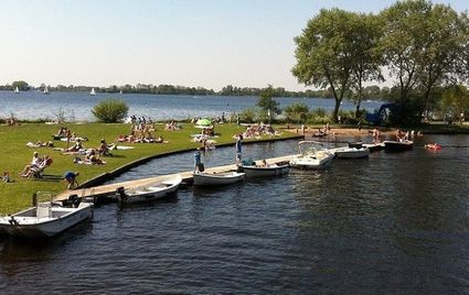 Mensen zonnen en ontspannen aan de oever van een meer met aangemeerde bootjes langs de steiger op een zonnige dag in het Groene Hart.