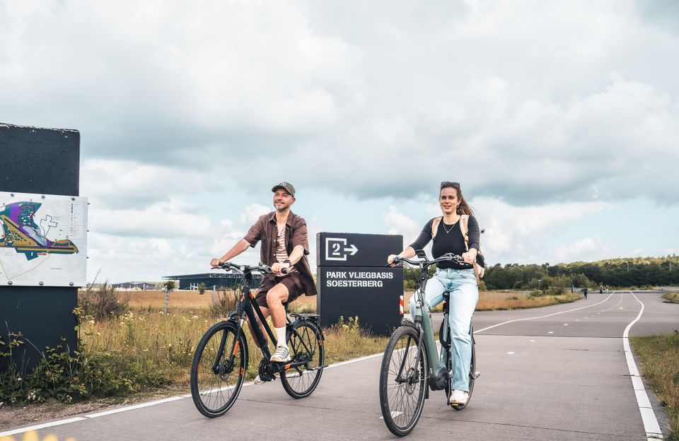 Twee fietsers rijden over de voormalige startbaan van Park Vliegbasis Soesterberg. Op de achtergrond ligt het Nationaal Militair Museum in het groene landschap van de Utrechtse Heuvelrug.
