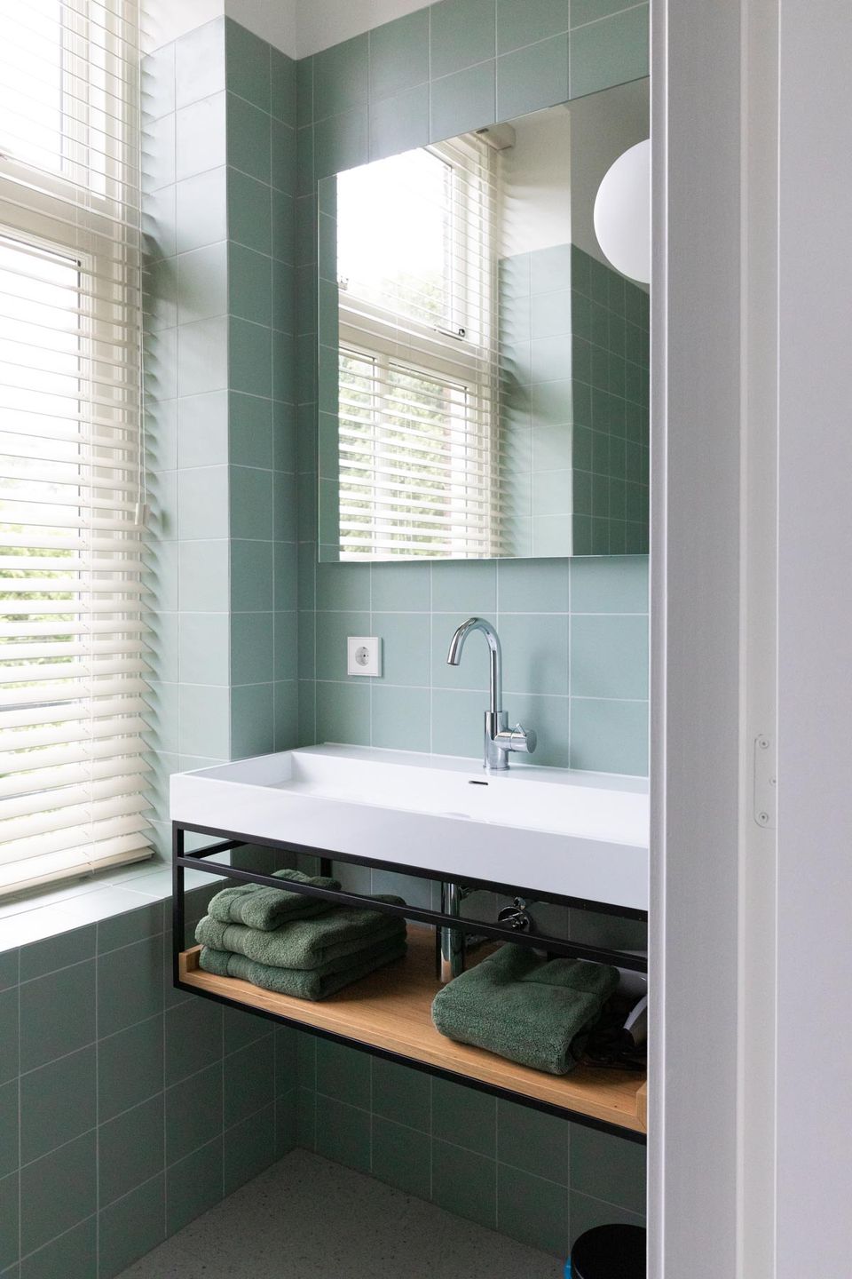 Badkamer van Boutique Pastorale in Montfoort met een moderne wastafel, groene wandtegels, grote spiegel en handdoeken op een open houten plank.