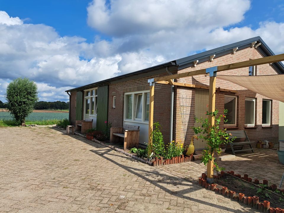 Bakstenen woning met groene luiken en houten terrasoverkapping met schaduwdoek, gelegen aan een erf naast landbouwgrond onder een blauwe lucht met wolken.
