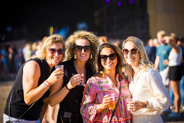 Vier lachende vrouwen met een drankje in de hand poseren op het zonnige festival ’t Wonder in Woerden, met een feestende menigte op de achtergrond.