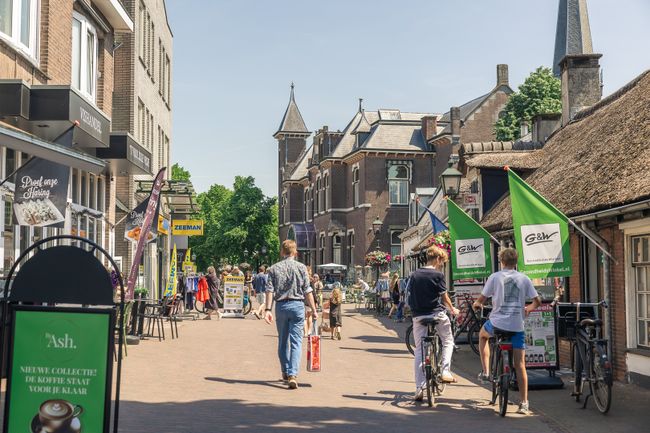 Bezoekers wandelen en fietsen door de levendige winkelstraat in het centrum van Baarn. Winkels, terrassen en historische bebouwing bepalen het straatbeeld.