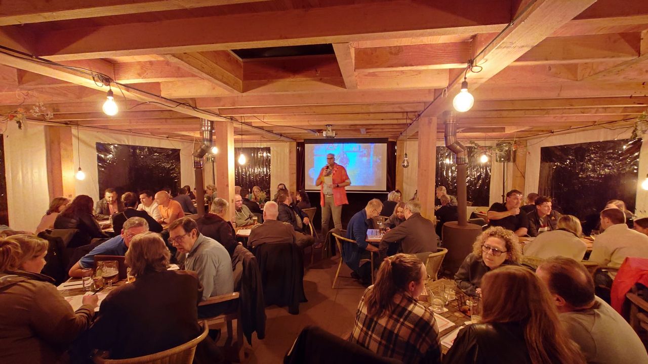 Foto van het interieur van het Boscafé De Rode Lelie met veel mensen zittend rond tafeltjes. In het midden staat de quizmaster voor een beeldscherm.