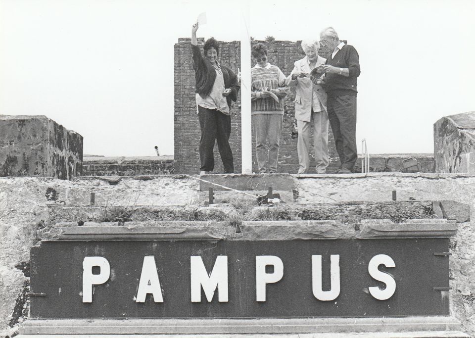 Vier mensen staan boven de tekst 'Pampus'bij de entree. Eén persoon zwaait vrolijk met een boekje naar de fotograaf, drie andere personen kijken aandachtig hun boekjes over Pampus door.