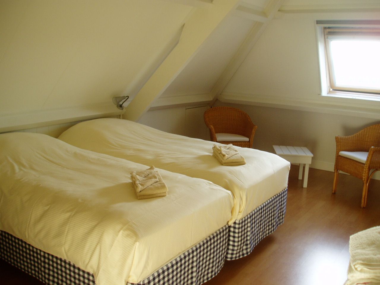 Slaapkamer onder de schuine kap met twee 1-persoonsbedden. Aan de rechterkant zit een dakraam met daaronder twee rotan stoelen en een tafeltje.