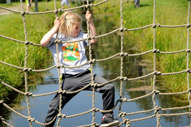 Poldersport Boerderij De Boerinn in Kamerik, Groene Hart. Kind klimt over een touwnet boven een sloot tijdens een actief kinderuitje, schoolreisje of familie-uitje in het landelijke poldergebied van Utrecht.