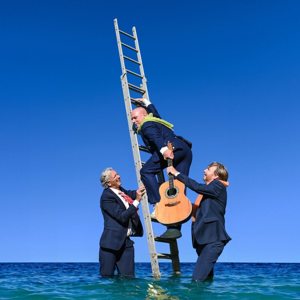 Drie mannen in donkerblauw pak. Twee van hen staan in het water en houden een ladder vast die ook in het water staat en daar klimt de derde man met gitaar op naar boven.