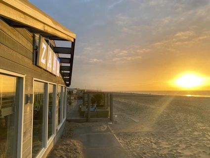 Uitzicht op zee met ondergaande zon, op het strand van Katwijk. Links is de voorkant en naam van strandpaviljoen ZAND zichtbaar, en een stukje van het terras.