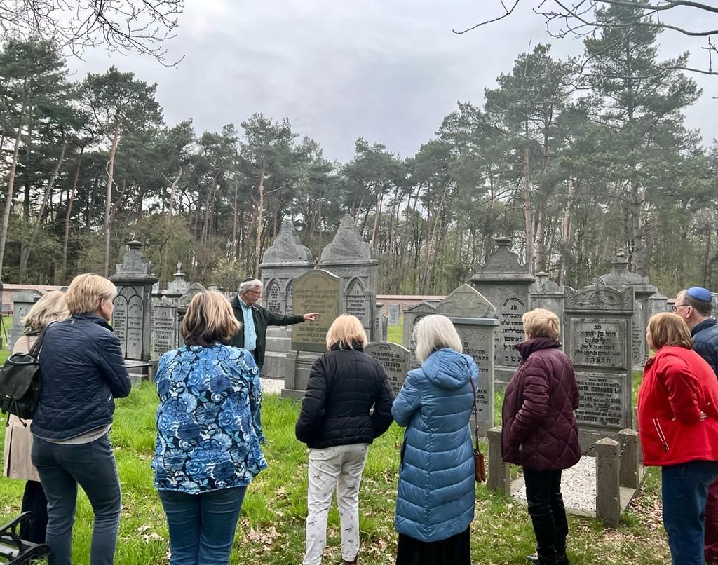 Rondleiding op de joodse begraafplaats. Een gids geeft uitleg aan een groep personen over de grafstenen. De graafplaats ligt in een groene omgeving met bomen op de achtergrond.