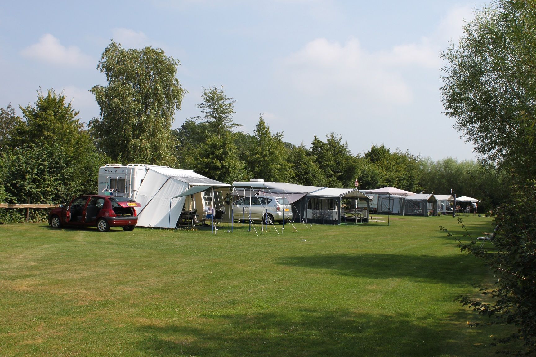 Een groen veld met diverse caravans met auto's en daarachter een rij grote bomen.