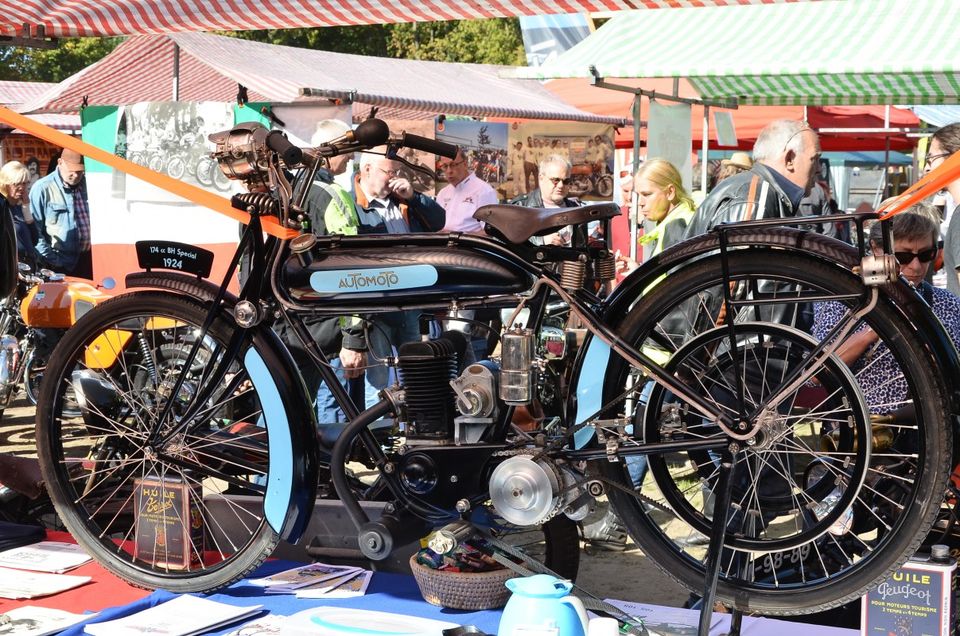 Historische motorfiets Automoto uit 1924 tijdens een oldtimerevenement in Woerden, Groene Hart, klassiek zwart model met lichtblauwe details gepresenteerd op een beursstand met bezoekers, marktkramen en vintage motoren op de achtergrond.