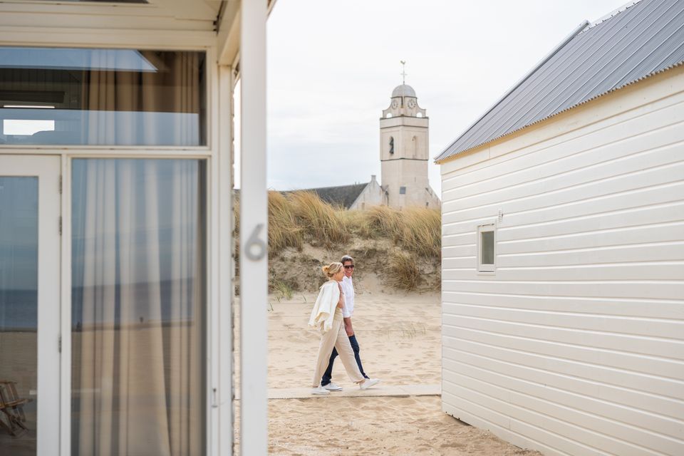 Foto tussen twee strandhuisjes door, van een wandelend koppel met de Andreaskerk op de achtergrond.