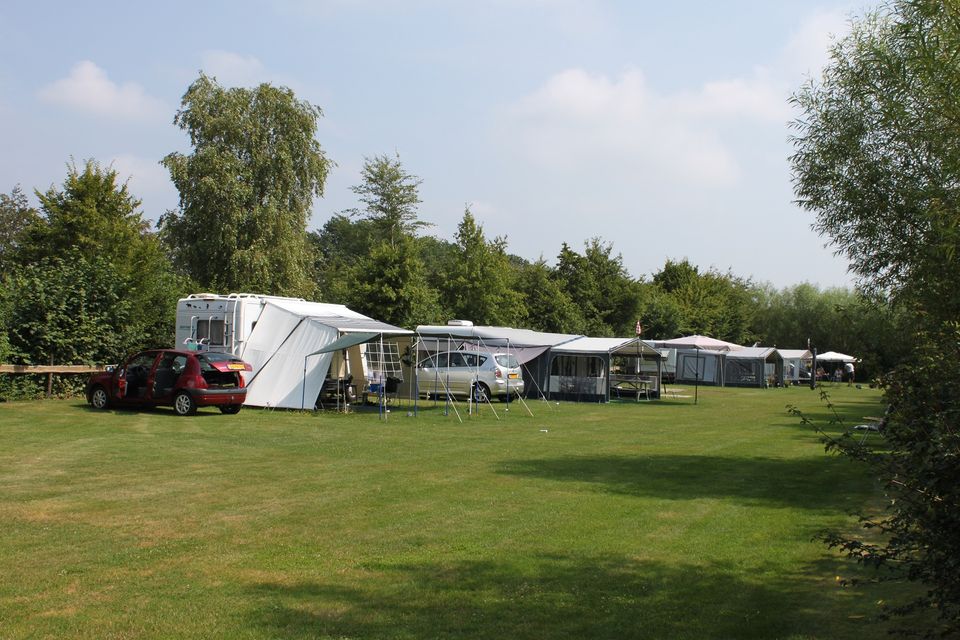 Een groen veld met diverse caravans met auto's en daarachter een rij grote bomen.