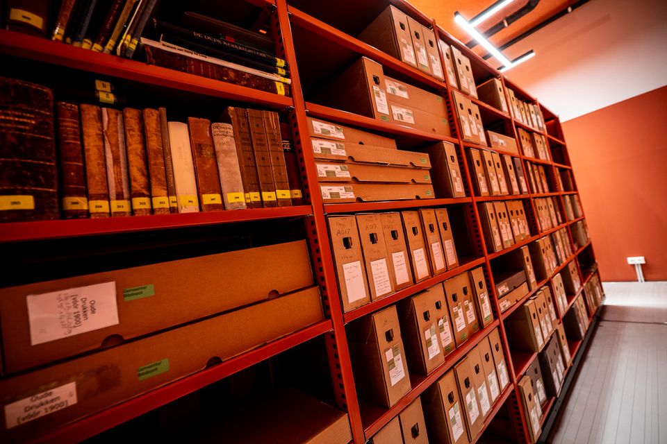 Rijen rode stellingen met archiefdozen en oude boeken in een archiefdepot, ordelijk gelabeld en opgeslagen voor bewaring en raadpleging.