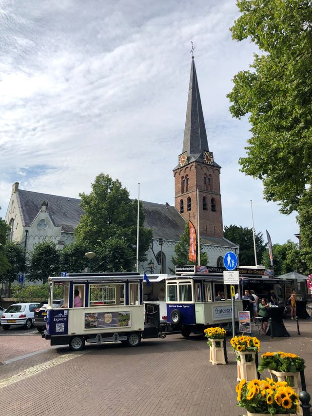 De blauw met witte Vorstelijk Baarn express die staat voor de Pauluskerk. Er staan bloemen op de voorgrond en er stappen mensen in.