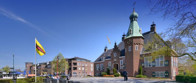 Gemeentehuis van Woudenberg aan het centrale plein, met vlaggen, zitgelegenheid en bebouwing van het dorpscentrum op de achtergrond. De foto laat het karakteristieke raadhuis en de centrale ligging in Woudenberg zien.