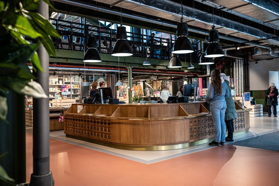 Bezoekers staan bij een houten servicebalie in een moderne bibliotheek, waar medewerkers helpen met vragen, onder industriële verlichting en met zicht op andere verdiepingen.