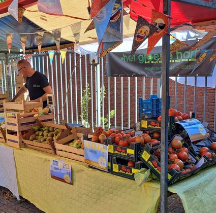 Evenement Roemeniemarkt Woerden, Groene Hart, groente- en fruitkraam met tomaten in kistjes, appels en peren op tafel met prijsaanduidingen en feestelijke vlaggetjes tijdens lokale goede doelenactie.