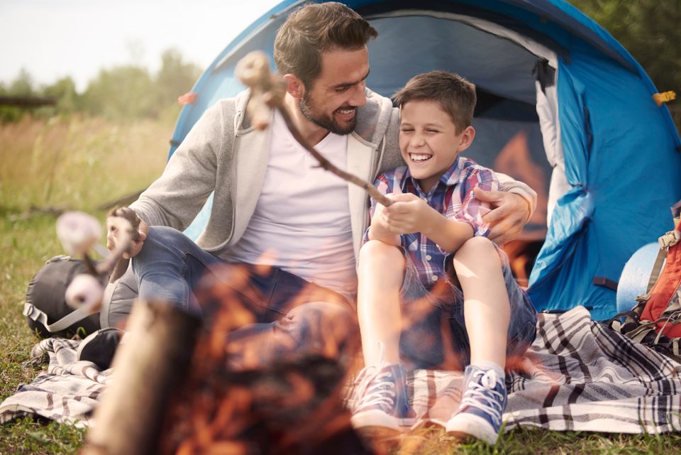 Een vader en een zoon zitten voor een tent marshmallows te roosteren boven een kampvuur.