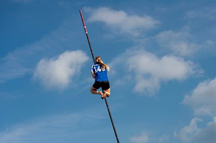 Jonge deelnemer aan het Nederlands Kampioenschap Polsstokverspringen klimt hoog in de polsstok tegen een blauwe lucht met wolken.