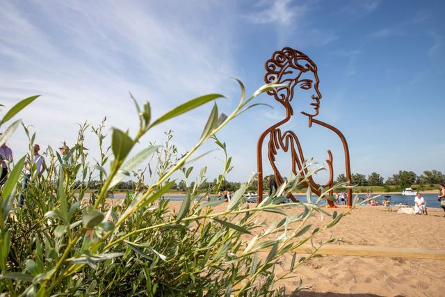 Op het strand van een recreatieplas staat een korten stalen beeld van een vrouw, met een hand voor de borst en met gekrulde haren.